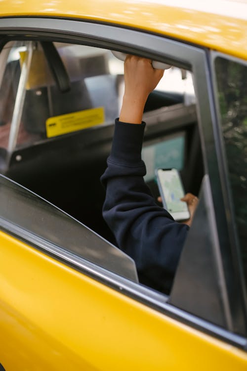 Free Unrecognizable passenger sitting on backseat and browsing smartphone while holding handle inside of yellow cab with lowered window during ride Stock Photo