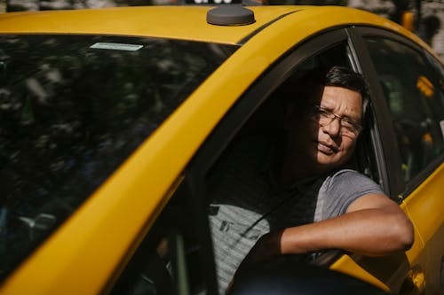 Adult male driver in glasses sitting in yellow cab and working while looking at camera