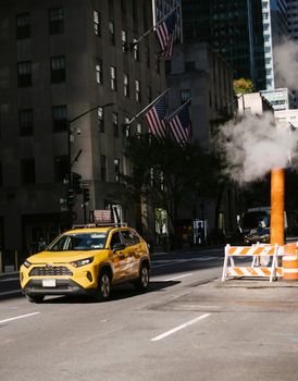 一辆黄色出租车驶过繁华的城市街道，街道附近冒着蒸汽