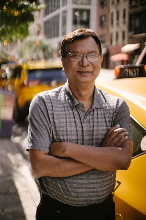 Smiling male driver in glasses and casual clothes standing on roadside with crossed arms and looking at camera