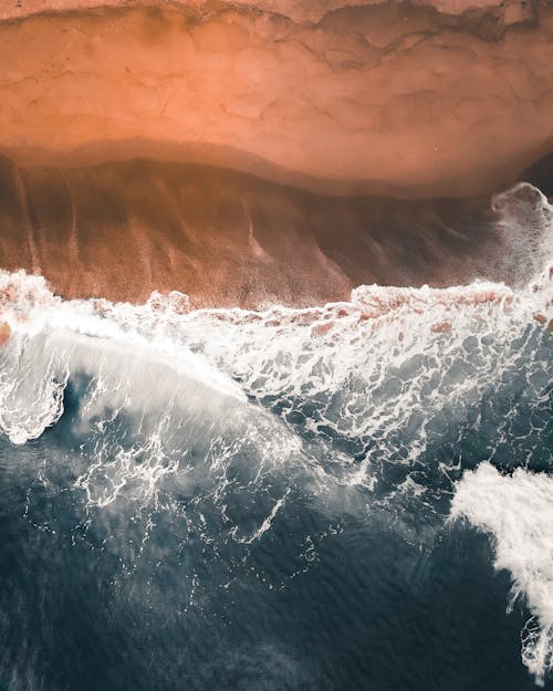 Aerial top view of powerful foamy ocean waving over desolated sandy beach on clear weather