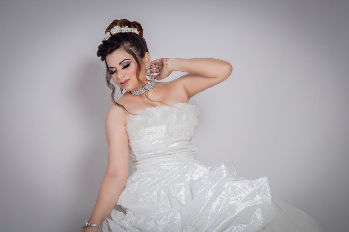 Female model dressed in bridal gown with makeup and jewelry touching neck gently standing against gray background