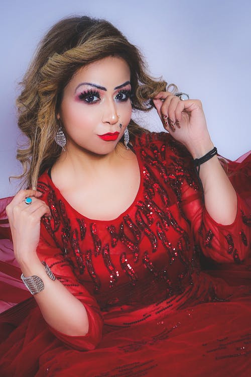 Young wavy haired female with bright makeup in red dress touching hair while looking at camera