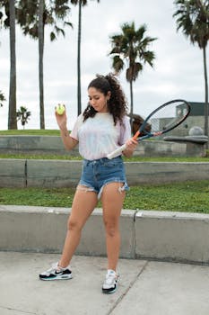 Female tennis player holding racket and ball in casual sportswear outdoors.