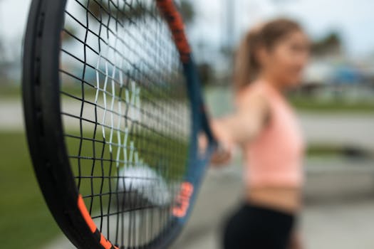 Close-up of a tennis racket with a blurred background featuring a woman, depicting athletic focus and motion.