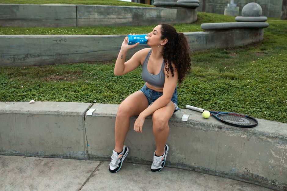 Young woman sitting on a bench with tennis racket and drinking water in a park.