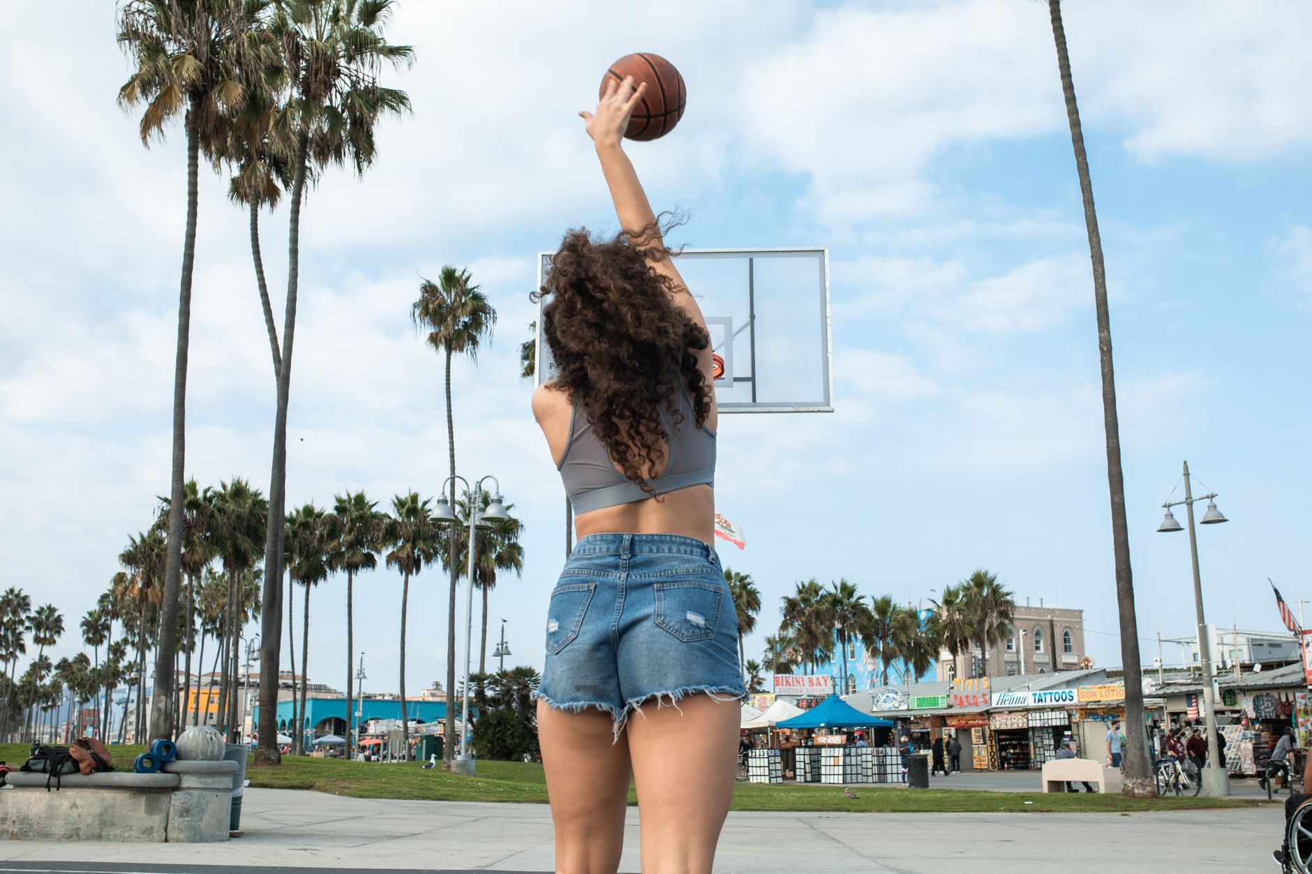 Woman playing basketball in a park with palm trees and blue sky.
