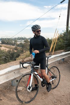 A cyclist in gear pauses with a drink beside his bike on a scenic road.