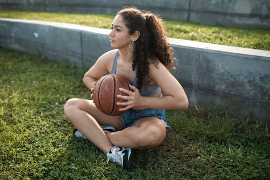 A young woman in casual sportswear sitting with a basketball on a grassy outdoor court, enjoying a sunny day.