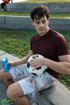 Man sitting outdoors with a soccer ball and holding a drink, enjoying leisure time.