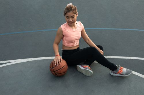 Free East Asian woman in sportswear sitting on an outdoor basketball court, holding a basketball. Stock Photo