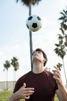 A young man skillfully balances a soccer ball on his head in a sunny, palm-filled park.