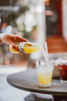 A hand pours citrus juice into a glass with ice and straw outdoors.