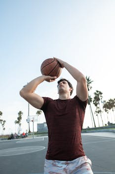 Man shooting basketball on outdoor court with palm trees in background.