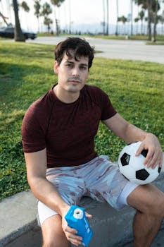 Caucasian man sitting outdoors with a soccer ball and drink in a sunny park.