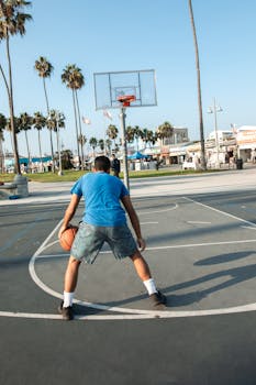 Teen dribbling basketball on outdoor court with palm trees and blue sky view.
