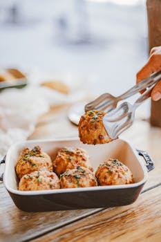Delicious homemade meatballs garnished with herbs, served with tongs in a ceramic dish.