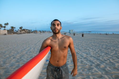 Free Man with surfboard standing shirtless on a sandy beach during dusk with a calm ocean backdrop. Stock Photo