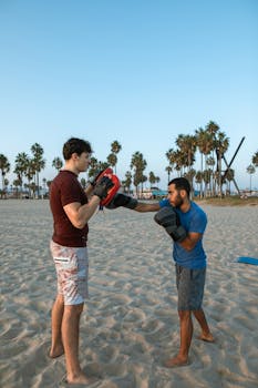 Two men engaged in a boxing workout on a sandy beach with palm trees around.