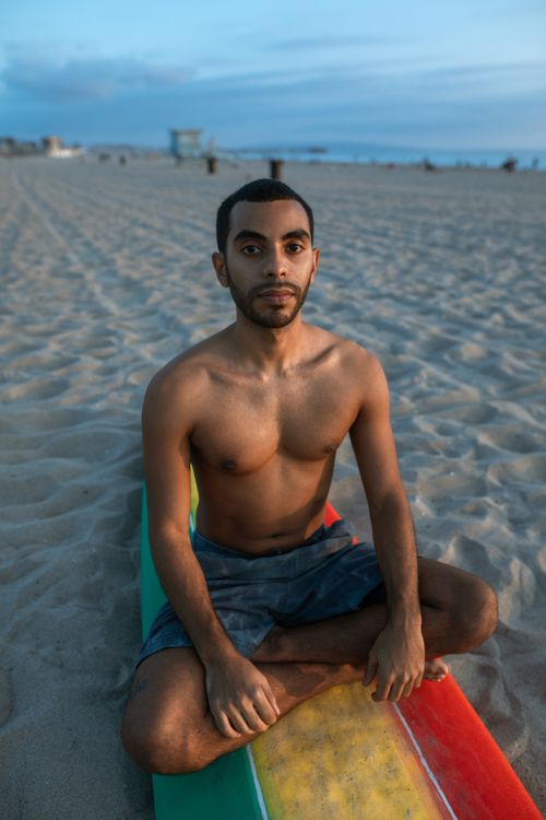 Free A shirtless man sits in meditation on a sandy beach with a colorful surfboard, creating a serene coastal vibe. Stock Photo