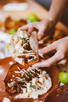 Close-up of hands holding a delicious taco with meat, creamy sauce, and a lime on the side.