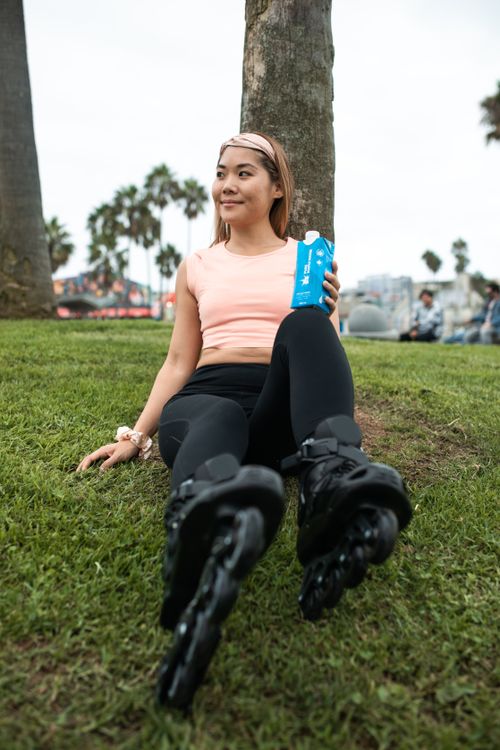 Free Asian woman in rollerblades resting on grass, enjoying a drink in the park. Stock Photo