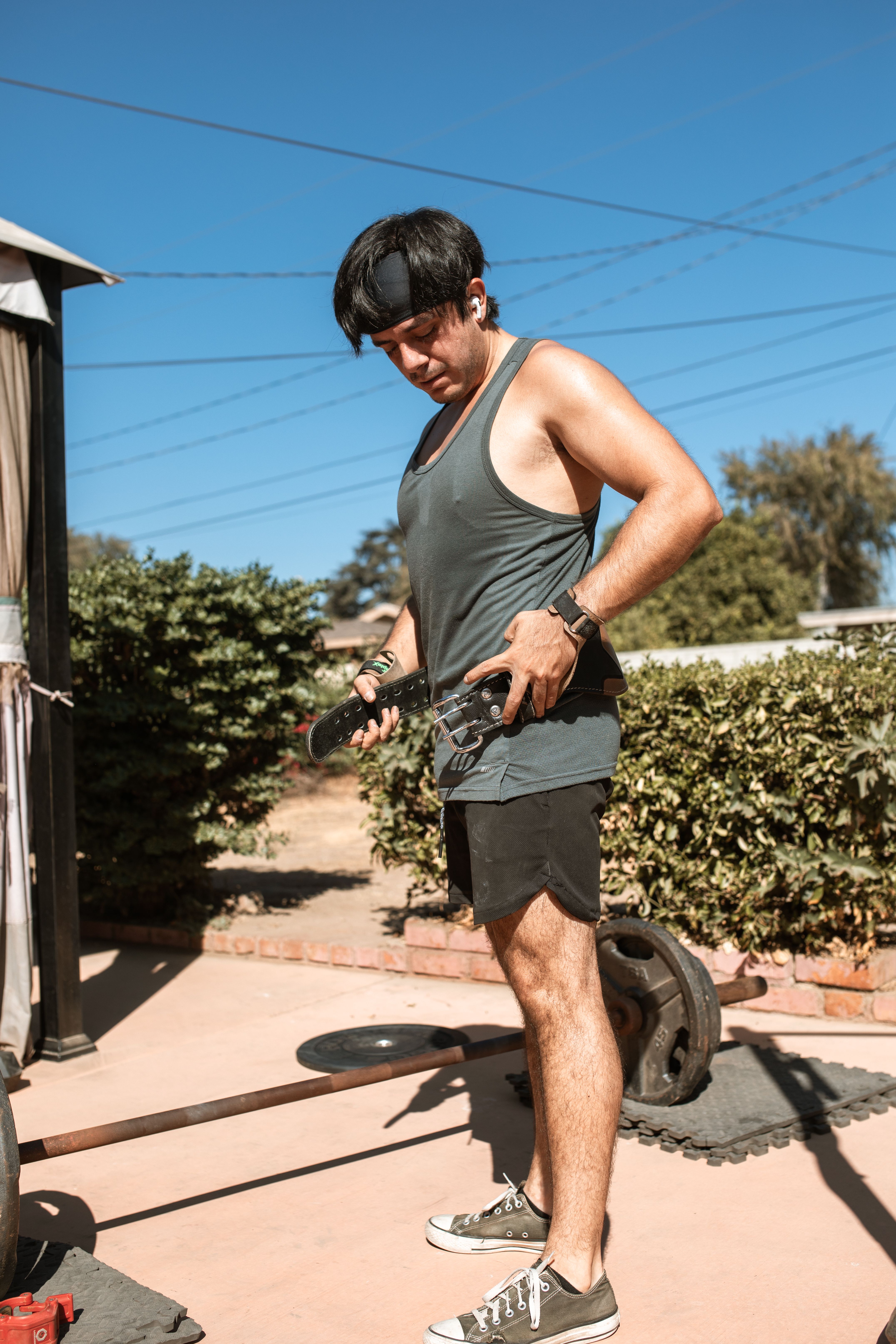 Man in tank top prepares for workout with barbell outdoors on sunny day.