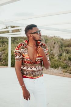 Confident young African American male traveler in stylish summer outfit and sunglasses relaxing in tropical resort and looking away pensively