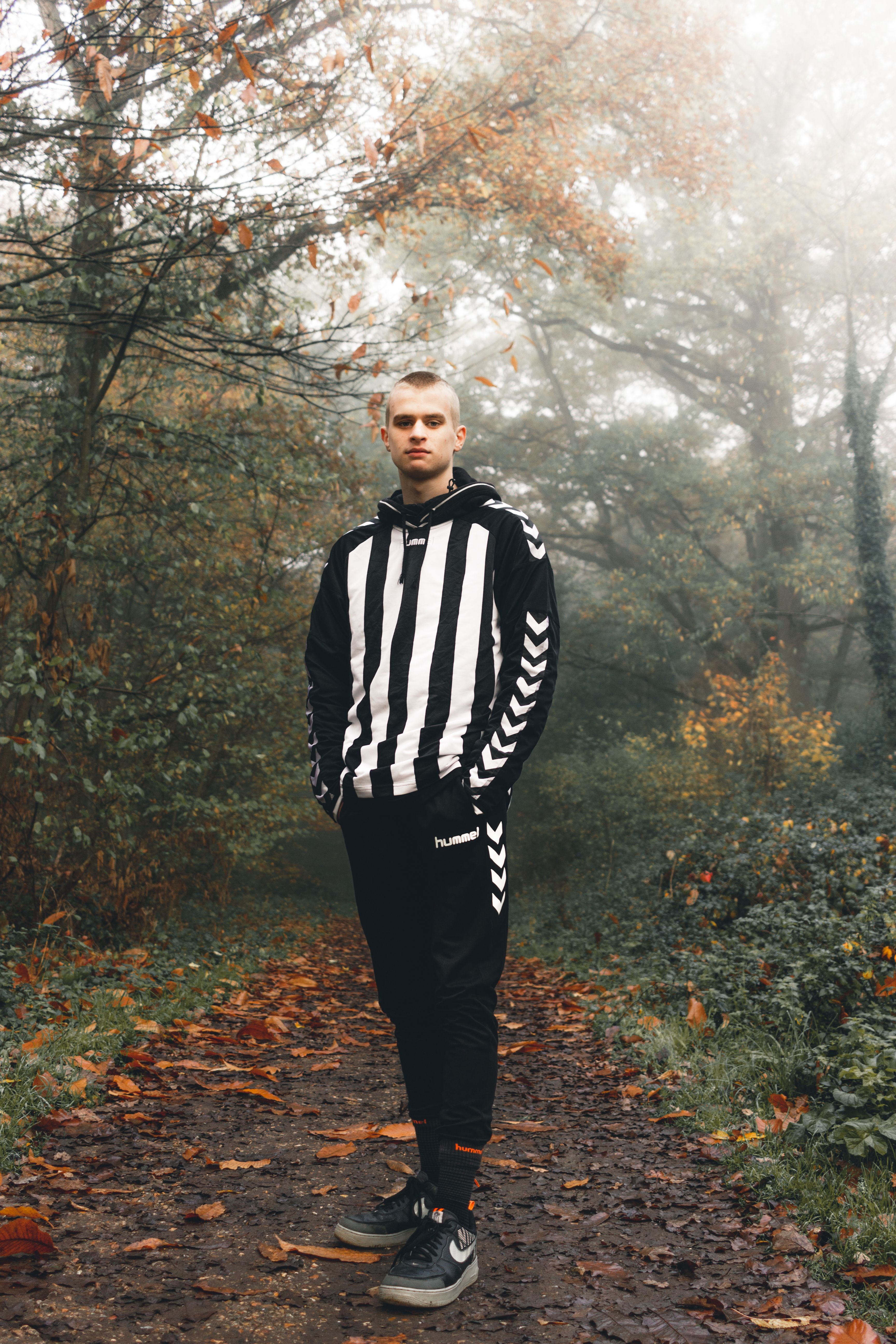 Young male in striped wear with hands in pockets standing on pathway in autumn woods and looking at camera