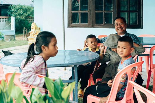 A family enjoying an outdoor gathering in Mukdahan, Thailand, with children and adults smiling around a table.
