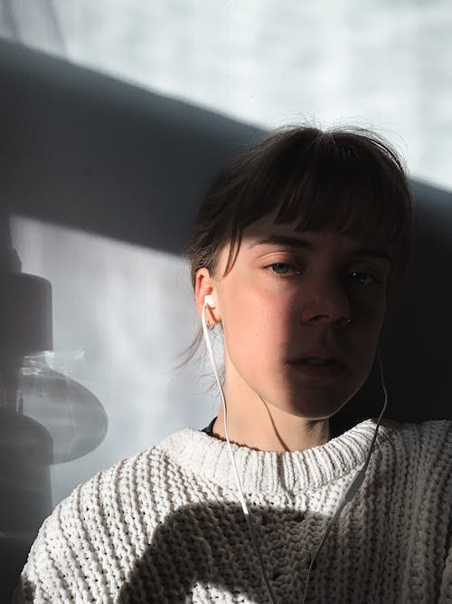 Melancholy female listening to music with earphones and looking at camera while sitting near wall with shadow on face in room with sunlight