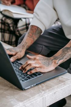 Crop faceless ethnic person with tattoos in casual clothes sitting at marble table and browsing netbook