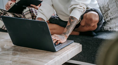 Crop anonymous tattooed ethnic person browsing internet on netbook while sitting with crossed legs on couch near friend in house