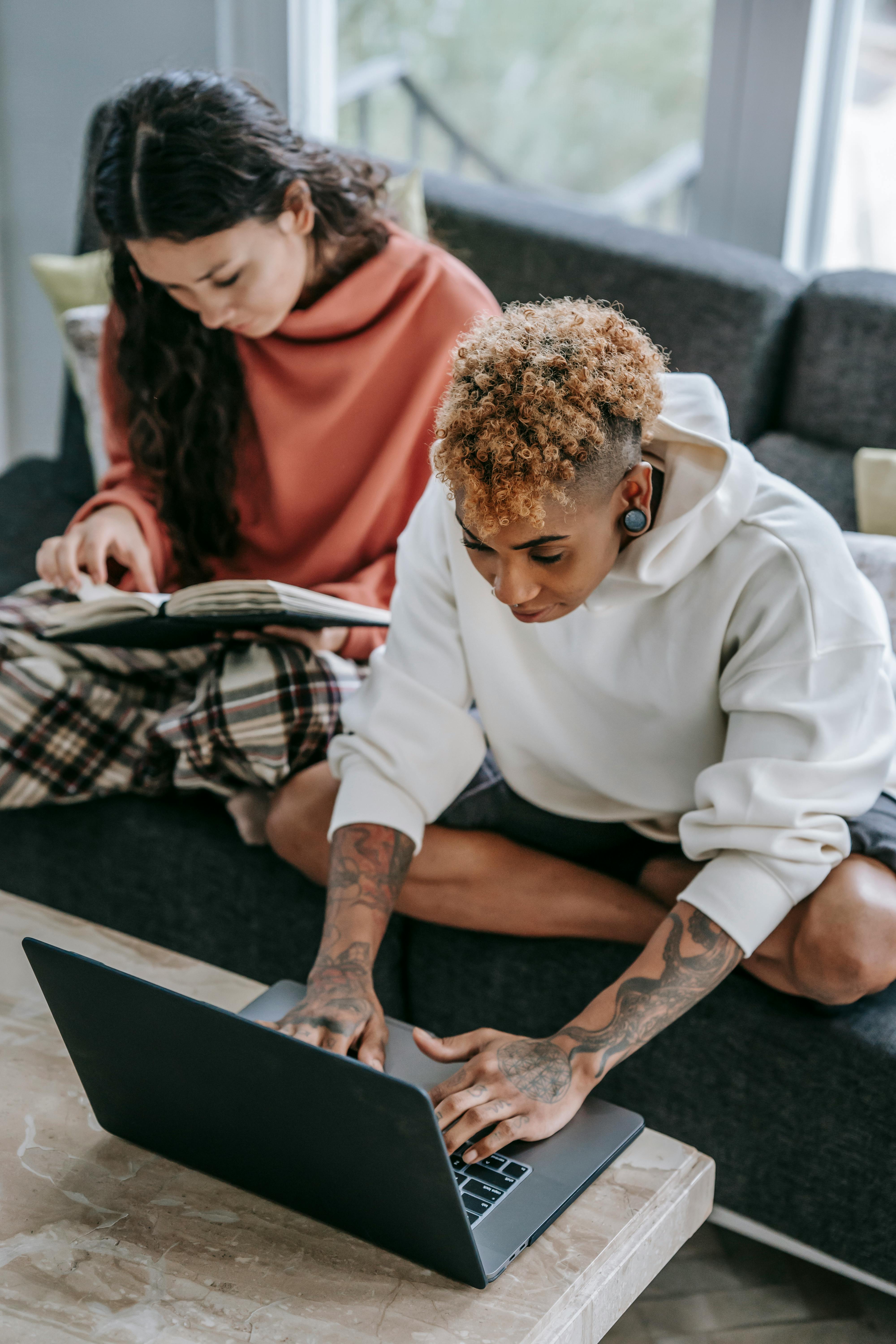 multiracial girlfriends typing on laptop and reading book on couch