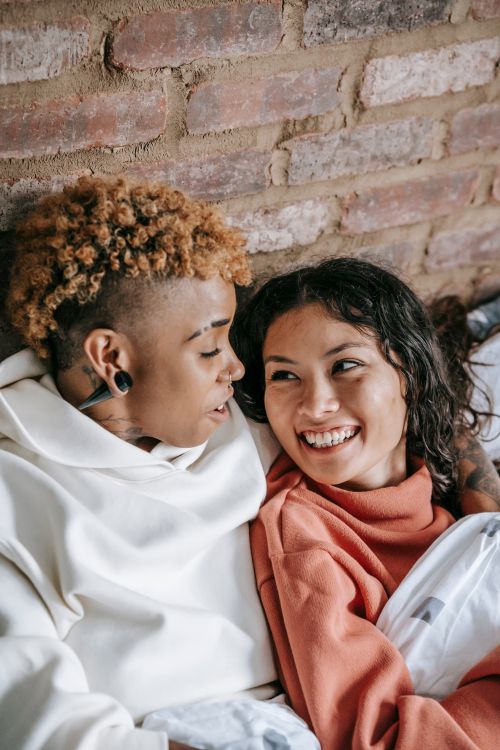 Free From above of cheerful young homosexual multiethnic girlfriends cuddling while talking in soft bed and looking at each other Stock Photo