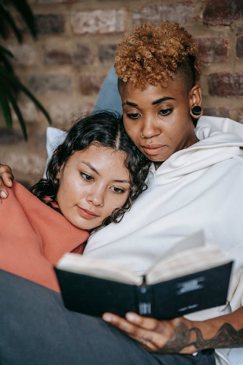 Homosexual multiethnic women reading textbook on bed at home