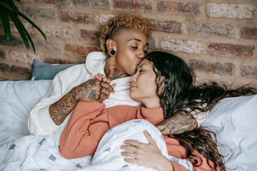 From above of young ethnic female with punk hairstyle embracing and holding hand of dreamy partner with closed eyes while resting in bed