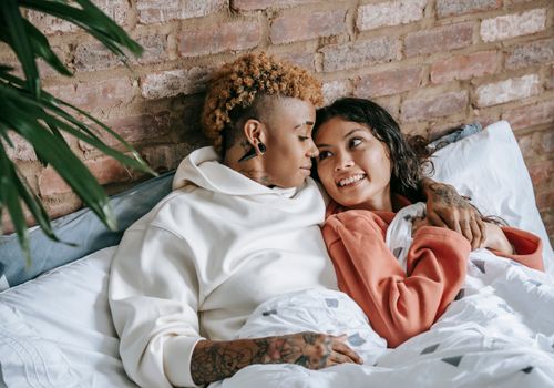 Free High angle of young content tattooed black woman embracing charming ethnic girlfriend while resting in bed and looking at each other Stock Photo