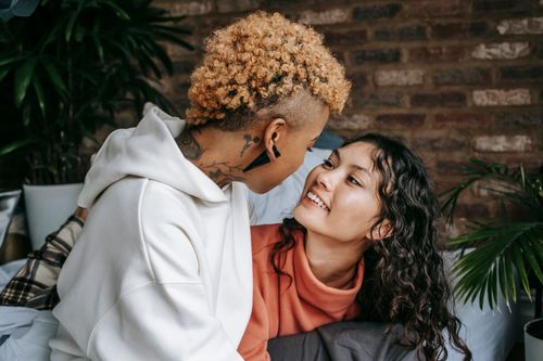 Free Anonymous tattooed ethnic woman with punk hairstyle sitting on bed with smiling girlfriend while looking at each other in house Stock Photo