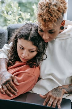 Crop tattooed black woman surfing internet on netbook while embracing homosexual ethnic girlfriend on couch in house