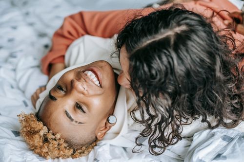 Free High angle of anonymous female cuddling cheerful homosexual ethnic partner while resting on cozy bed at home Stock Photo