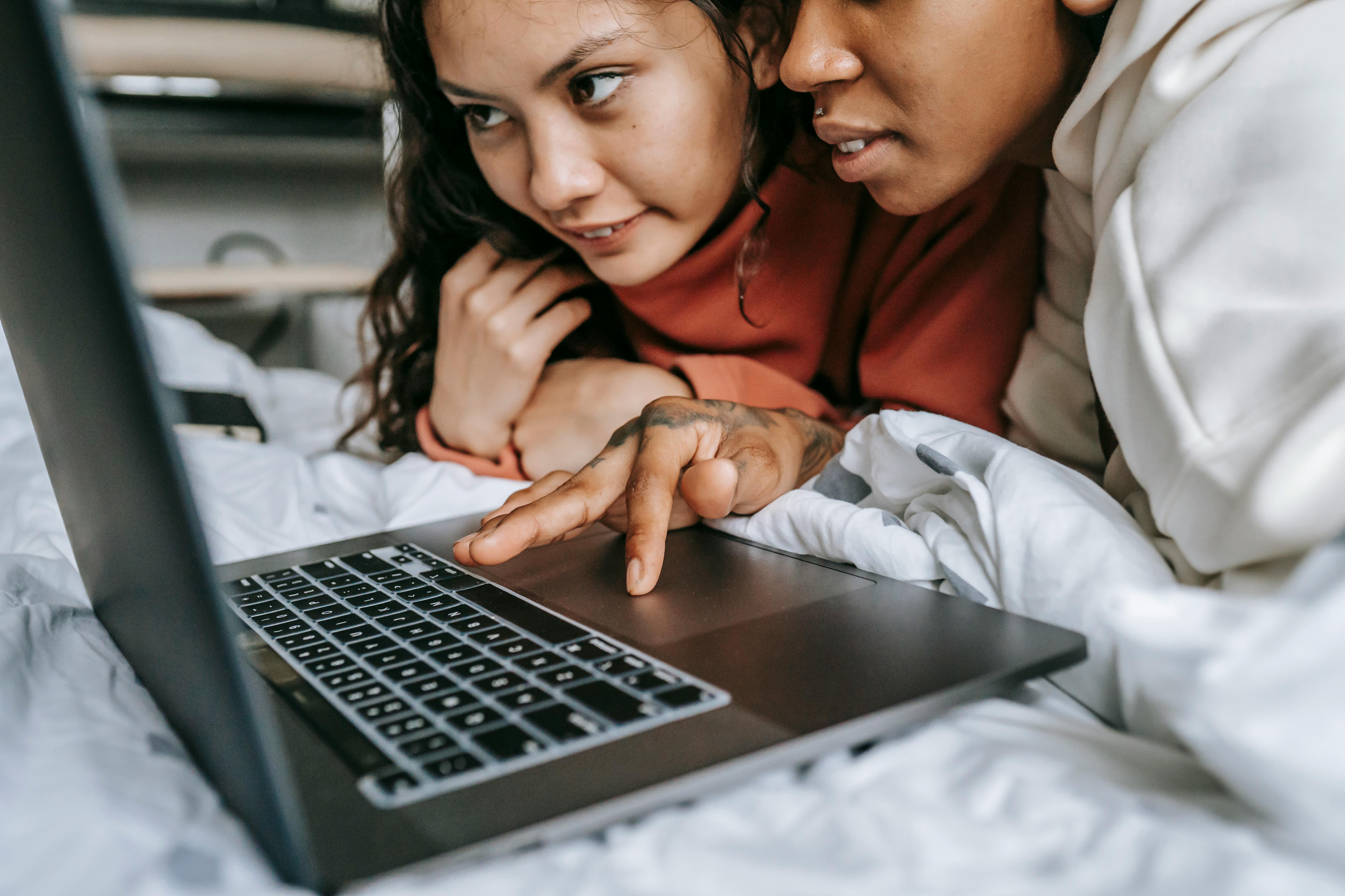 crop multiracial girlfriends browsing laptop on bed