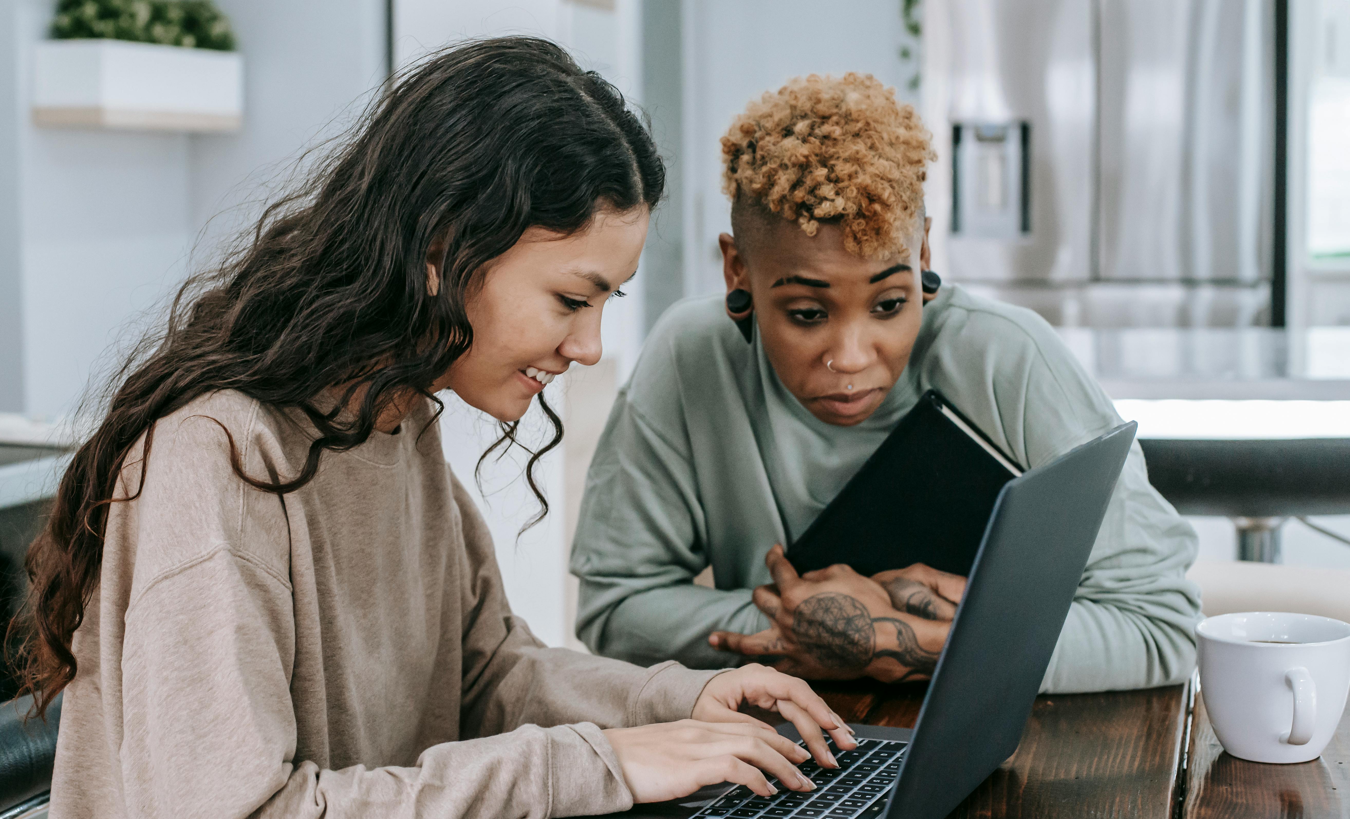 Two diverse women sharing a happy moment while using a laptop indoors. Smiling and engaged.