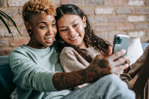 Happy African American female taking photo with smiling Hispanic female while resting together on comfortable couch