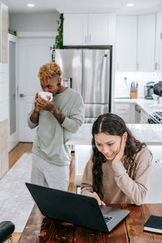 Two young women in a cozy kitchen enjoying coffee and working on a laptop, capturing a relaxed morning vibe.