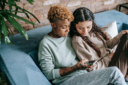 Concentrated young African American female scrolling feed in social network while resting on couch with interested Hispanic female