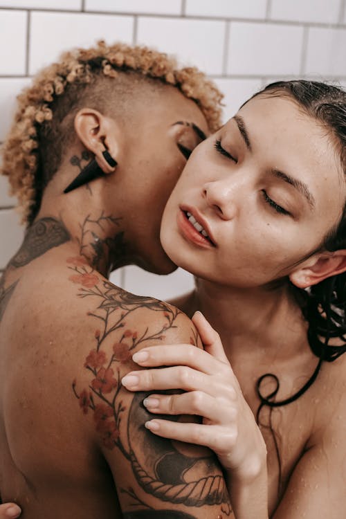 Side view of nude lesbian multiracial couple standing close and kissing while hugging under shower stream