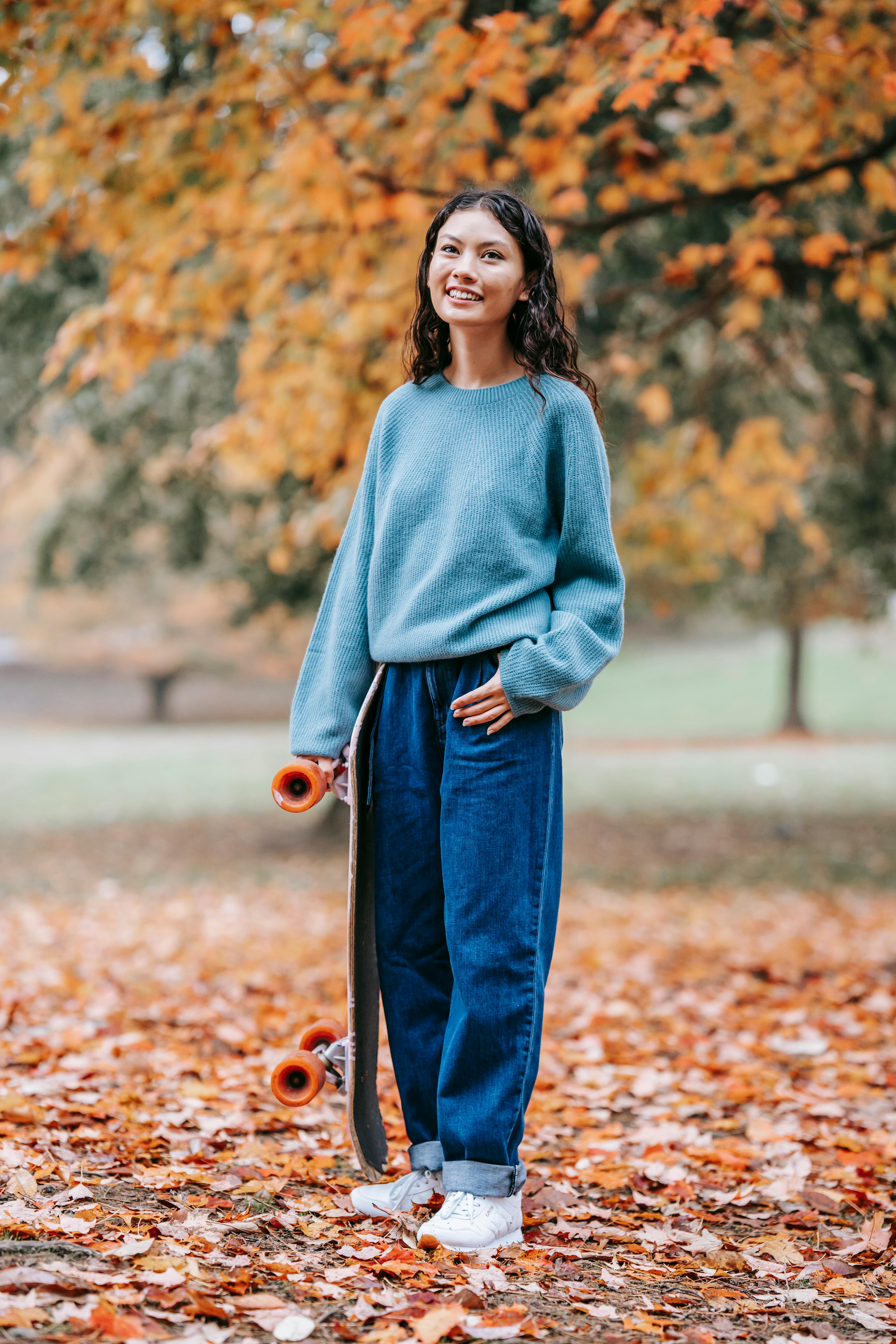 Full length of smiling Hispanic female in casual outfit standing in park in autumn with skateboard