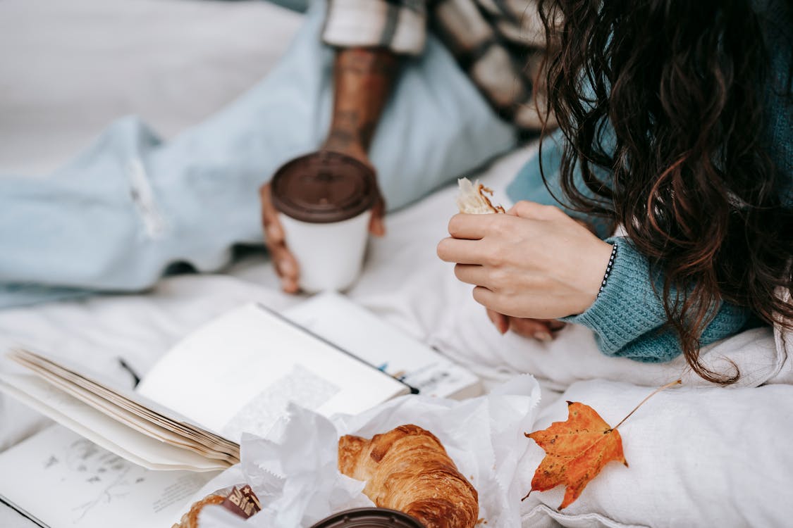 Unrecognizable multiracial couple with takeaway hot drink and croissant lying on white plaid with opened book during picnic in autumn day