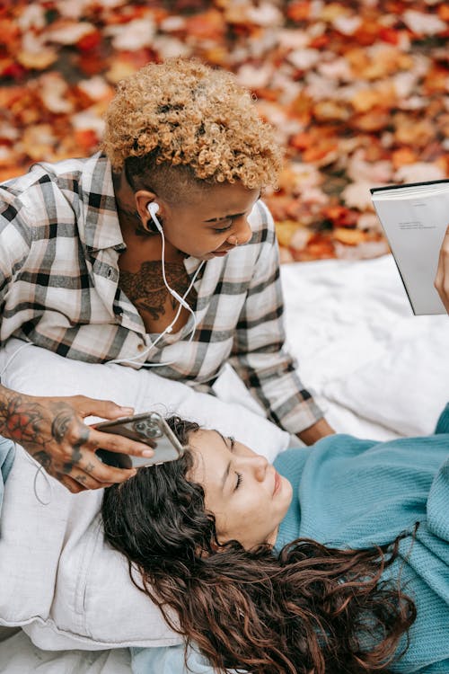 From above of Hispanic woman with book lying on blanket near African American girlfriend with smartphone and earphones with fallen leaves on blurred background
